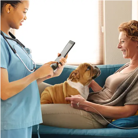 A doctor in scrubs wearing a stethoscope reads from a tablet device while talking to a patient laying on a comfortable couch petting a dog while receiving an IV infusion.