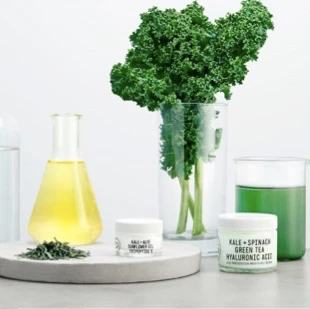 Youth to the People product containers with beakers of plant extracts on a white granite table top with kale leaves and a small pile of herbs.