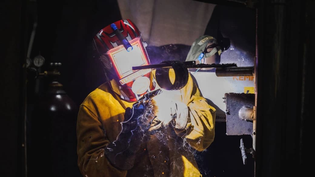 Two welders in protective helmets and gear welding a metal component as sparks fly