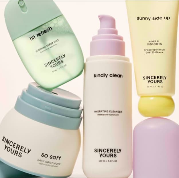 Collection of Sincerely Yours skincare products including a green hit refresh serum mist, white so soft daily moisturizer jar with teal lid, kindly clean hydrating cleanser pump bottle, yellow sunny side up mineral sunscreen tube, and a partial view of a purple product, all arranged against a light background